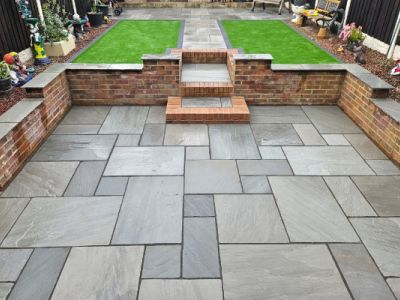 A backyard patio with large gray stone pavers is shown. The patio is bordered by brick walls and has a small section of green artificial grass with a brick pathway leading to it.
