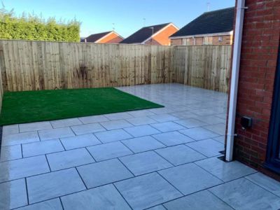 A newly paved patio surrounds a rectangular patch of artificial grass in a backyard. The patio is made of light grey rectangular tiles laid in a staggered pattern.
