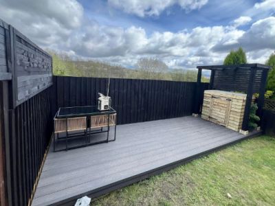 A modern backyard deck features a dining set and a wooden storage unit under a pergola. The area is enclosed by black wooden fences.