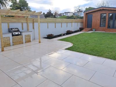 An outdoor patio area features a built-in grill station and a wooden garden room. The patio leads to a lush green lawn bordered by a white wall.