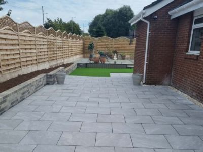 A newly paved patio extends from a brick house towards a wooden fence. The patio is made of large, light gray rectangular pavers.
