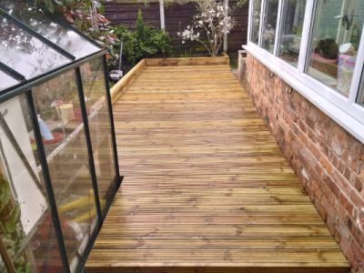 A wooden deck runs alongside a brick wall with a greenhouse on the left and a sunroom on the right. The deck is made of wooden planks and appears to be recently installed.