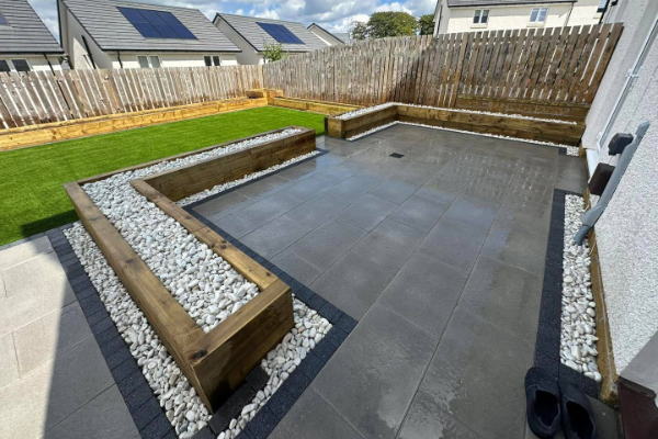 a modern backyard patio and lawn with wooden planter boxes filled with white stones. The patio features large grey tiles and is bordered by dark paving stones.