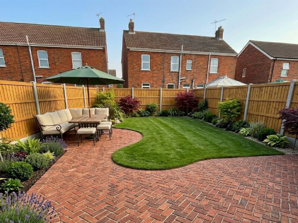 A well-maintained backyard features a brick patio with outdoor seating under an umbrella, a lush green lawn, and diverse landscaping. The scene is framed by wooden fencing and bordered by residential houses.