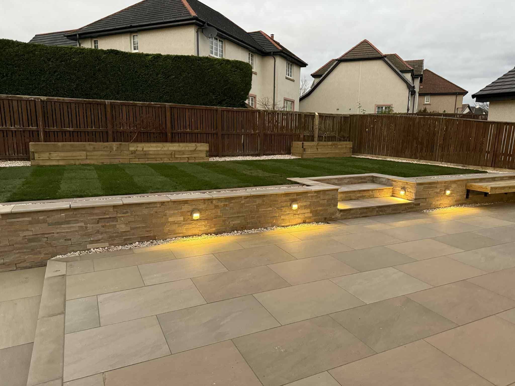 A well-lit, modern backyard patio with tiered landscaping and built-in seating. The image showcases a neat lawn, stone walls with integrated lighting, and a paved patio area.
