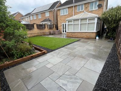 A newly paved patio with a grass area and garden beds is visible in the backyard of a brick house. The patio features large, light gray flagstones laid in a staggered pattern, bordered by black gravel.