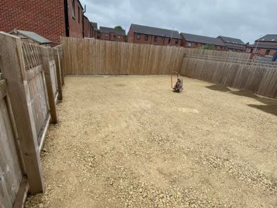 A backyard is being prepared with a layer of gravel and a compactor machine is present. The area is enclosed by wooden fences and residential buildings are visible in the background.