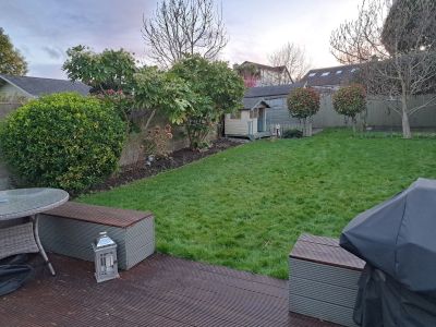 A backyard features a lawn, a small shed, and various trees and bushes. A wooden deck with seating and a covered grill is in the foreground.