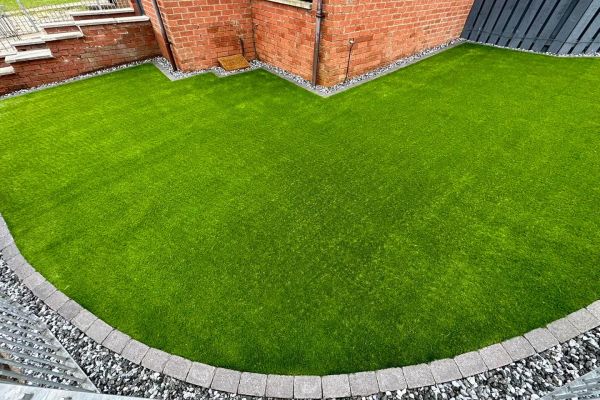 A vibrant green artificial lawn is bordered by grey paving stones and dark grey gravel. The lawn is situated next to a red brick building with a dark fence.